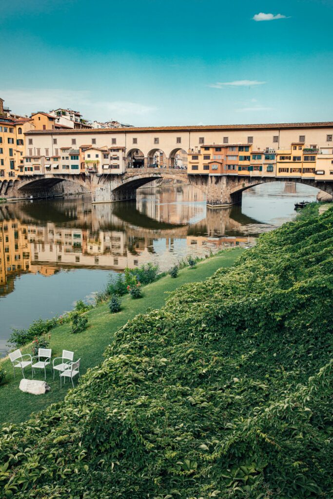 A scenic view of the Ponte Vecchio in Florence, Italy, reflecting in the Arno River, with lush green grass and chairs in the foreground under a clear blue sky.