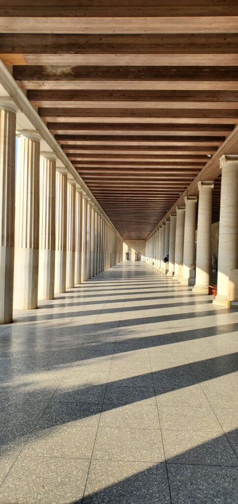 Interior view of a long corridor featuring tall columns and wooden beams, with sunlight casting shadows on the polished floor in Athens, Greece. 