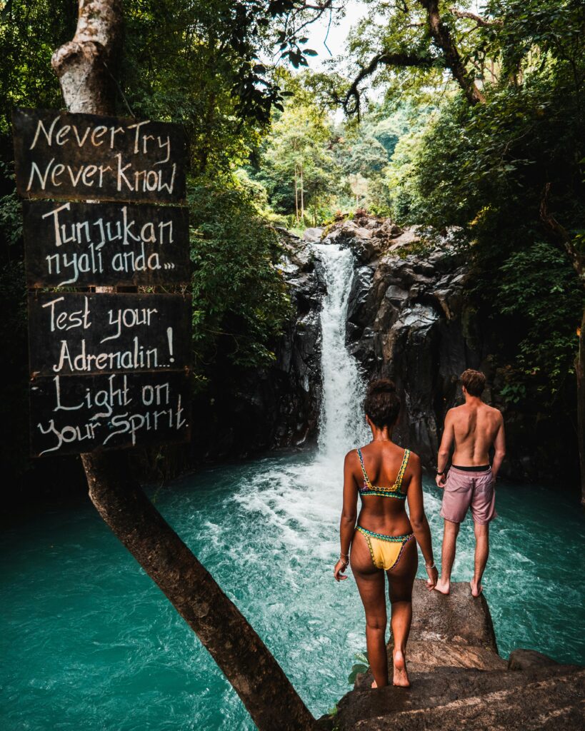 A woman in a bikini and a man stand on a rock near a waterfall surrounded by lush greenery, with a sign promoting adventure on a personalized travel experience. 
