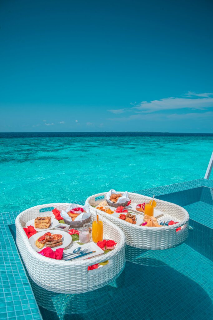 Two wicker trays filled with breakfast items, including fruits and pastries, resting on the edge of a swimming pool with clear blue water in the background through tailored travel experiences and bookings.