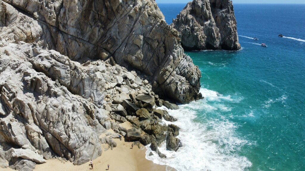 Aerial view of rugged cliffs and turquoise ocean waters at Cabo San Lucas, with a sandy beach and small figures of people enjoying the shore.