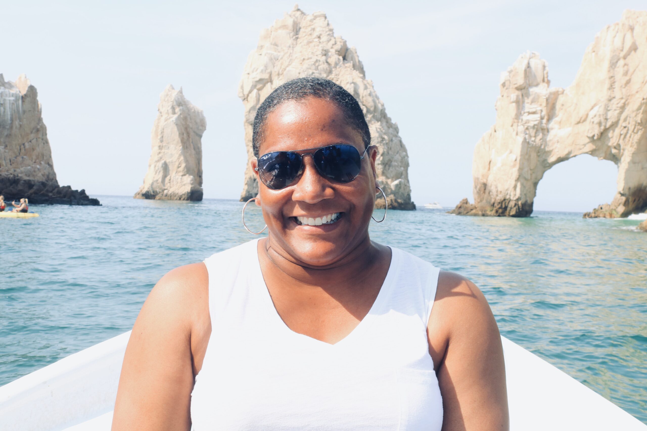 Founder woman wearing sunglasses, posing on a boat with rocky formations and calm ocean waters in the background. With Emerald Dreamscape vacations, personalized travel experiences are available. These are tailor-made for you. 